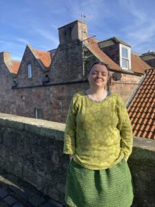 A photograph of Abigail Thomas who is wearing a lime green jumper with flower pattern. She is standing on a roof top with a red brick building in the background