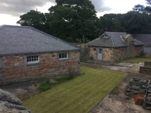 External image of two stone buildings that will be used as studios/workshops. There is grass and concrete paving. There are large trees in the background