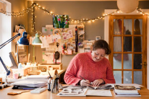 Illustrator Ali Edwards in her studio. She is wearing a pink jumper and in there is festoon lights in the background